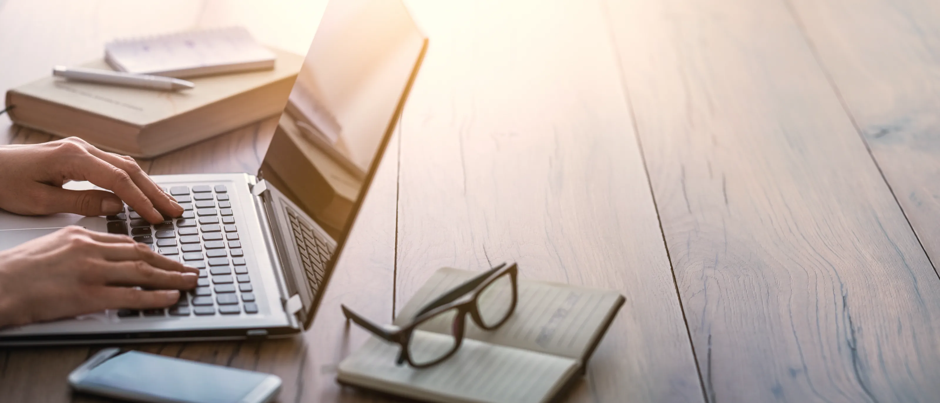 Hands typing on a laptop on a wooden desk, with an open notebook, reading glasses and warm afternoon window light